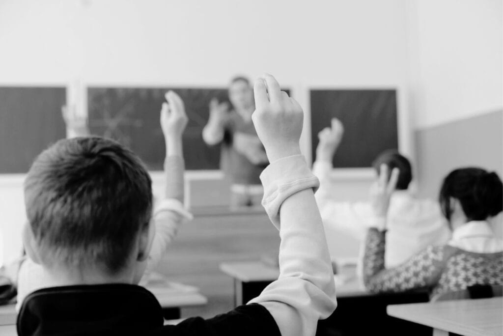 Home 1 Children sitting in a conventional school classroom lifting hands.